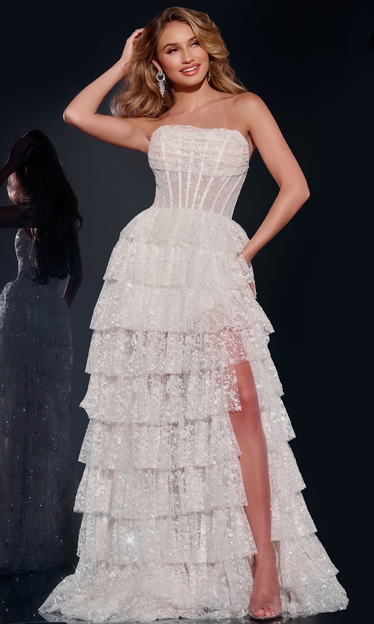 A woman stands smiling against a dark background in the Jovani 40031 strapless sequin prom ball gown, featuring a tiered white design and high slit.